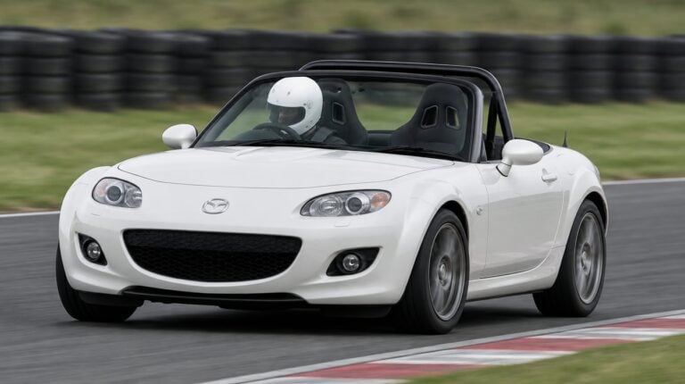 A white Mazda MX-5 Mk3 NC cornering on a race circuit with a helmeted driver sitting low in a black bucket seat
