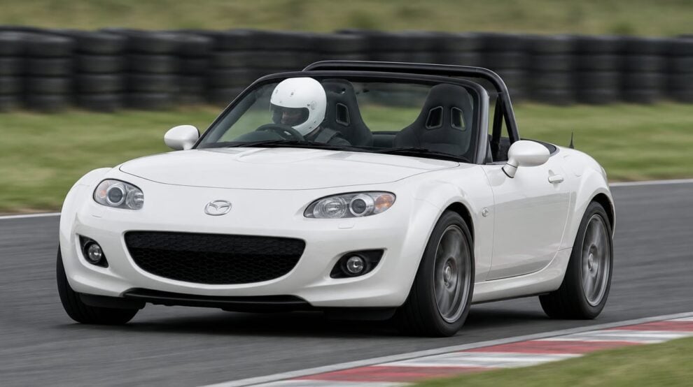 A white Mazda MX-5 Mk3 NC cornering on a race circuit with a helmeted driver sitting low in a black bucket seat
