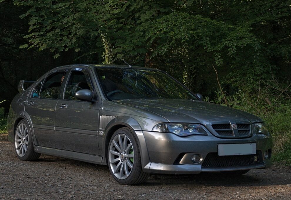 A 2005 MG ZS+ 110 saloon in XPower Grey parked on a woodland track, wearing green brake calipers and multi spoke alloys