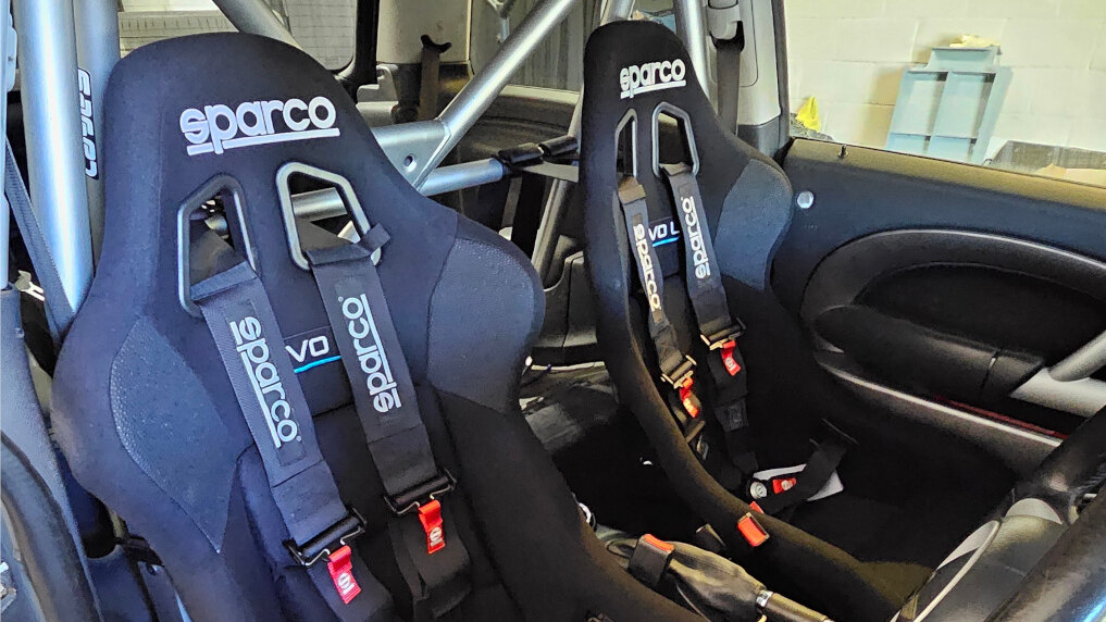 Two Sparco Evo L bucket seats with black Sparco four-point harnesses in a Mini R53, with a half cage behind