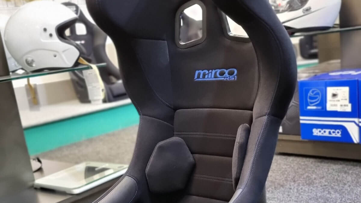 A black Mirco RS1 FIA bucket seat with blue embroidery on the showroom floor, with helmets on glass shelves behind