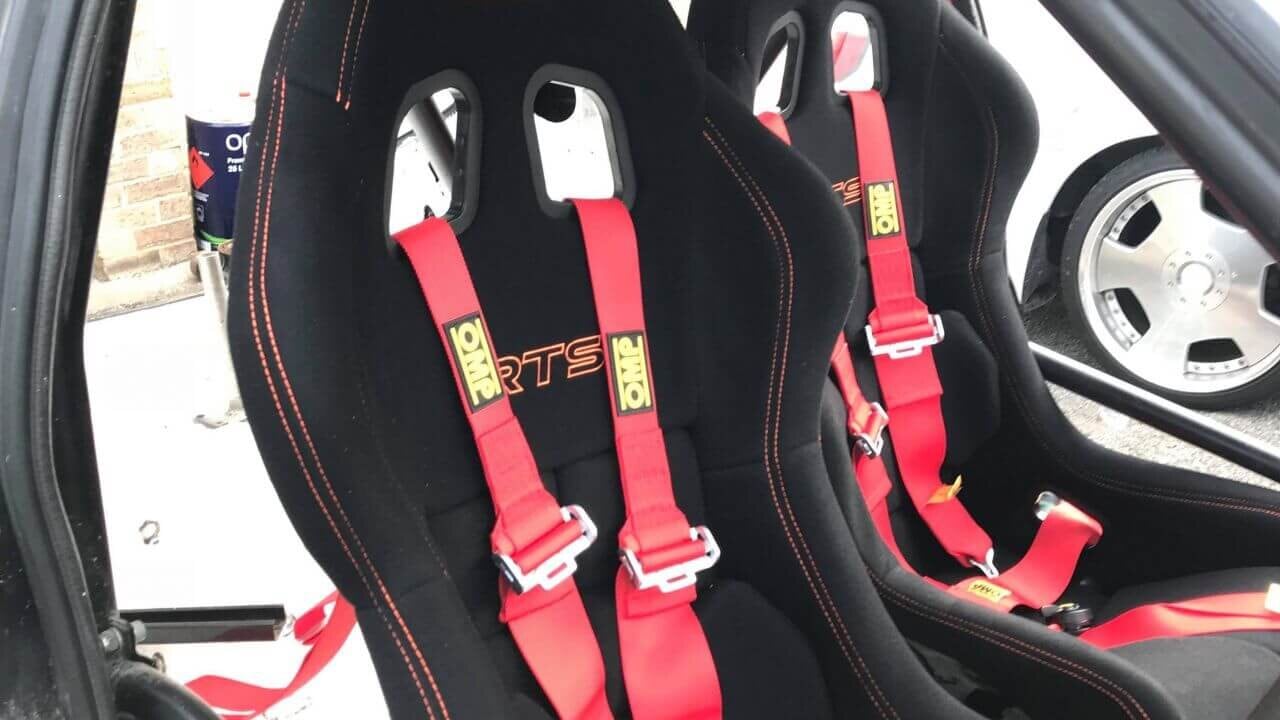Two black Mirco RTS bucket seats with orange stitching and red OMP harnesses in a caged Peugeot 205