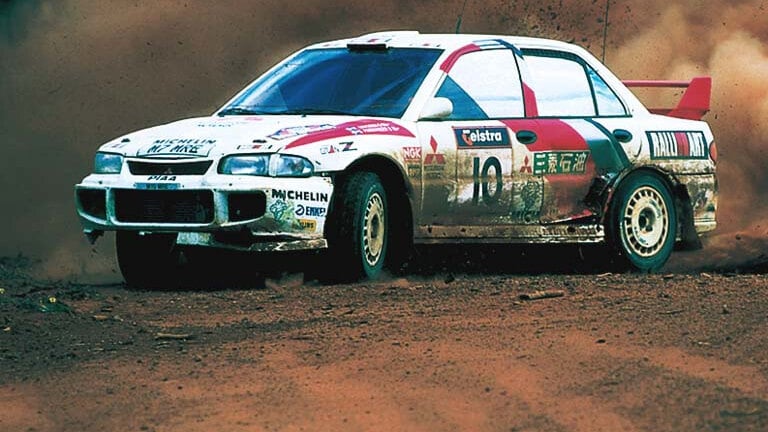 A white Mitsubishi Lancer Evolution rally car in Ralliart livery kicking up dust on a gravel stage