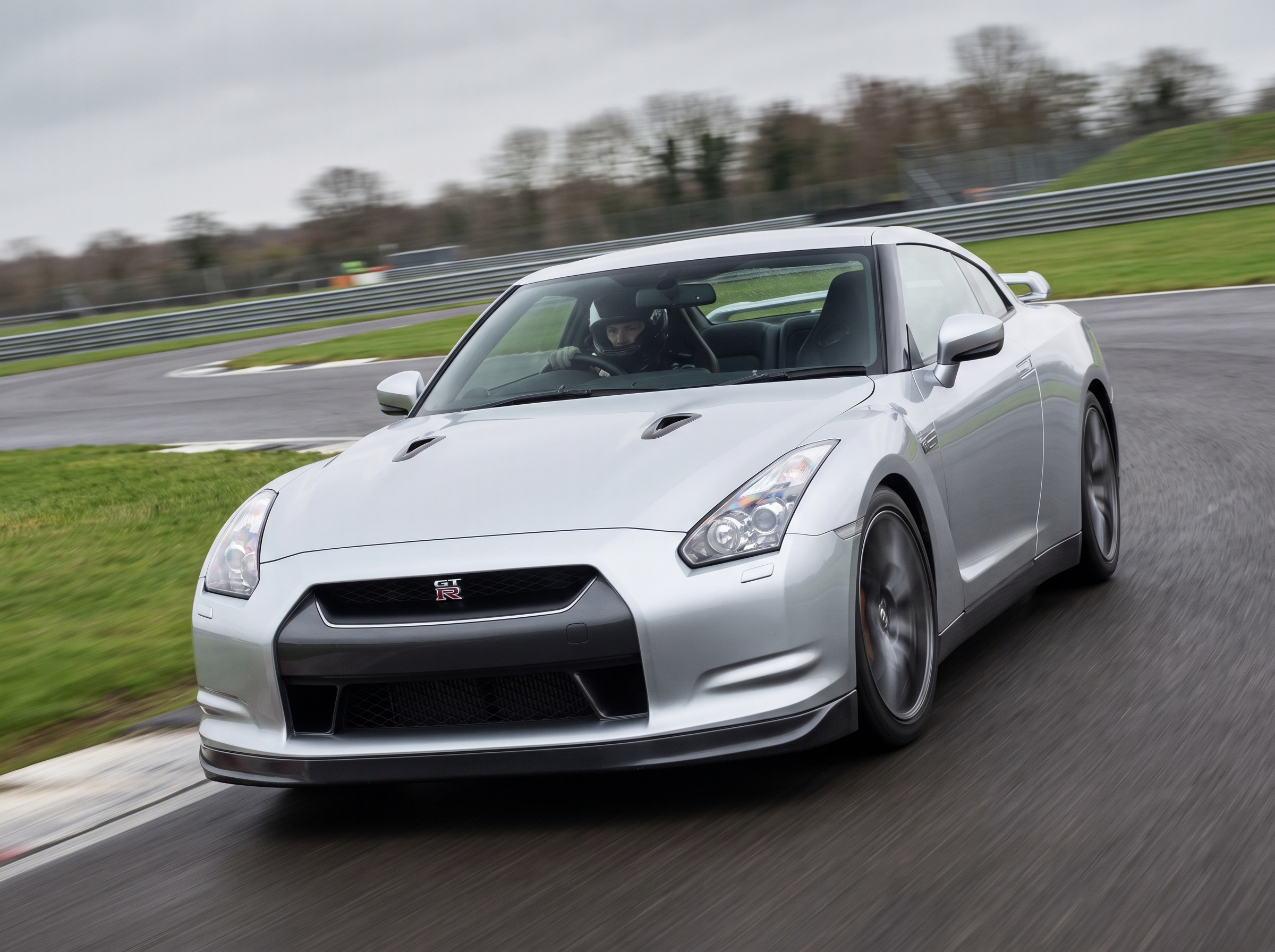 Silver Nissan GT-R R35 cornering on a British race circuit with helmeted driver at the wheel