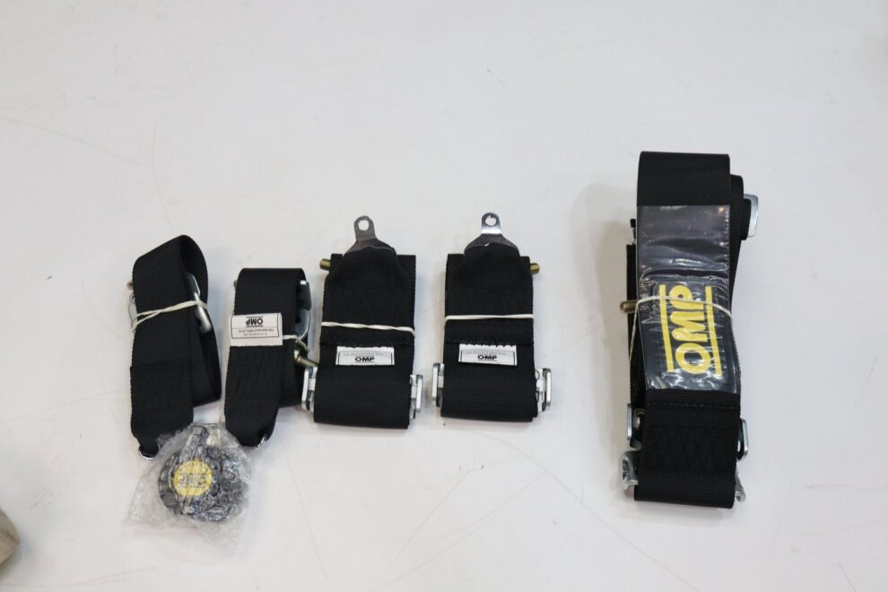 The parts of a black OMP six point harness laid out on a workshop floor: shoulder straps, lap straps, sub straps and the rotary buckle