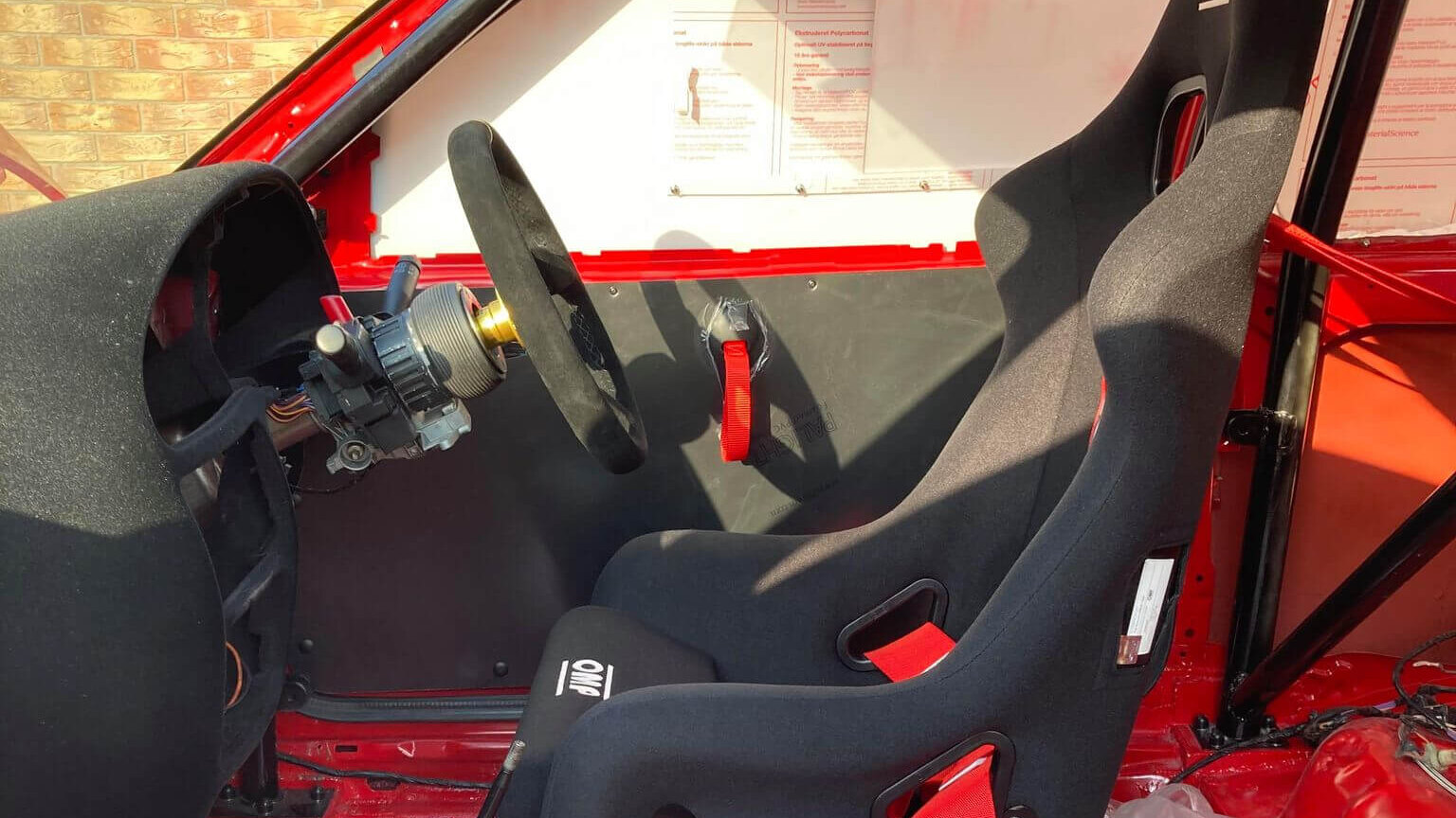 An OMP First R bucket seat with red harness beside a suede OMP steering wheel in a stripped red caged car
