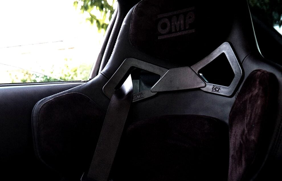 Close view of an OMP RT FIA bucket seat fitted in a Nissan 350Z, showing the embroidered OMP panel at the top of the backrest, the alcantara shoulder pads, the moulded harness slot trim and the car standard three point seat belt running across the seat