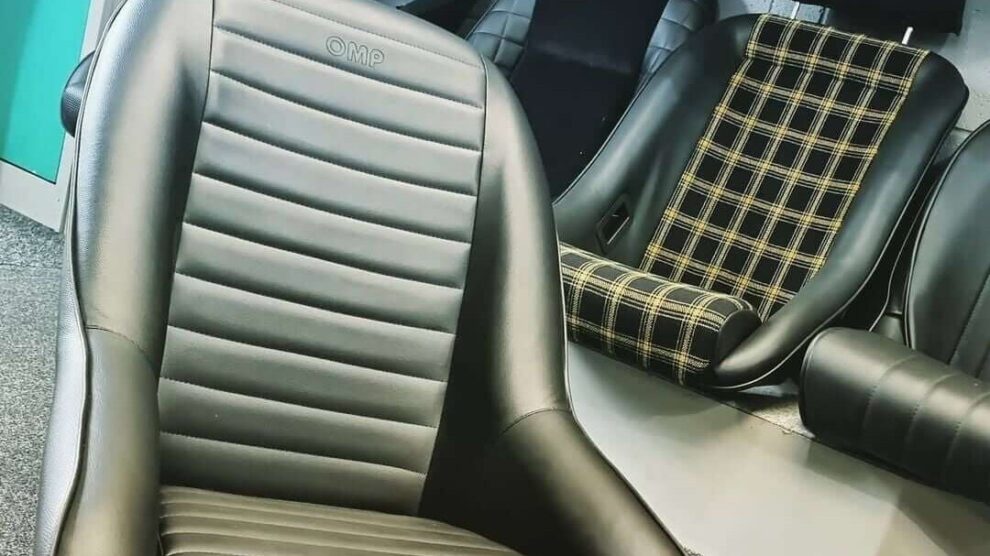 A black OMP Silverstone classic bucket seat with horizontal ribbing in the showroom, with classic seats including a tartan one on the floor behind