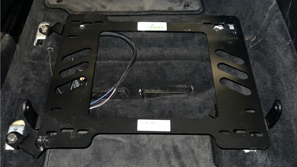 A black Planted Technology seat bracket bolted to the floor of a BMW E39 5 Series, with the carpet and seat wiring beneath it
