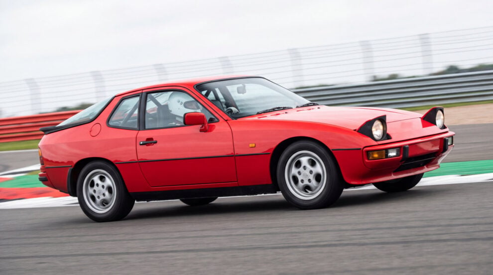 Red Porsche 924 with a roll cage visible through the side glass, cornering on a British race circuit with a helmeted driver at the wheel