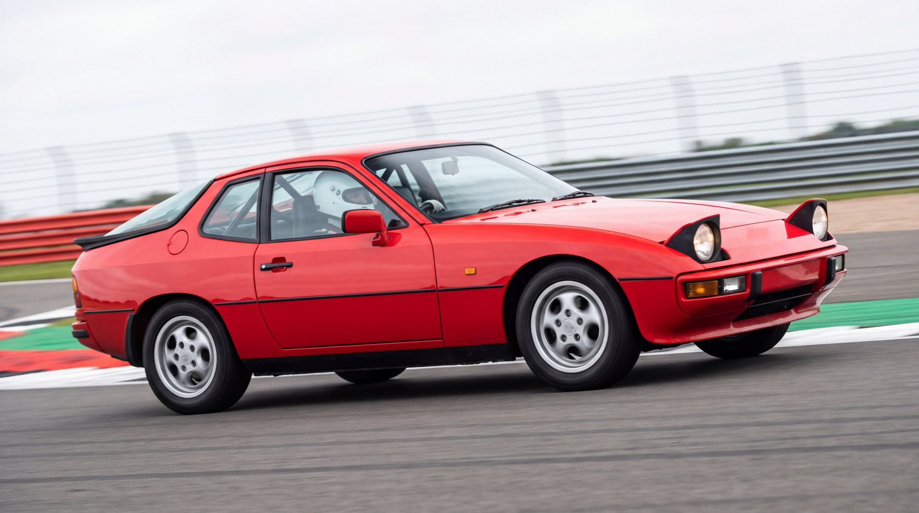 Red Porsche 924 with a roll cage visible through the side glass, cornering on a British race circuit with a helmeted driver at the wheel