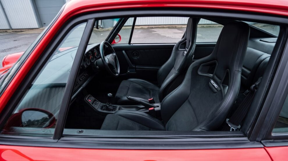 Looking down into the cabin of a red Porsche 964 through the open window of a shut door, showing a pair of Recaro Sportster CS reclining seats facing forward in black leather with suede centre panels, the Recaro wordmark on the shoulder bolster and the classic five dial 911 dashboard ahead of them