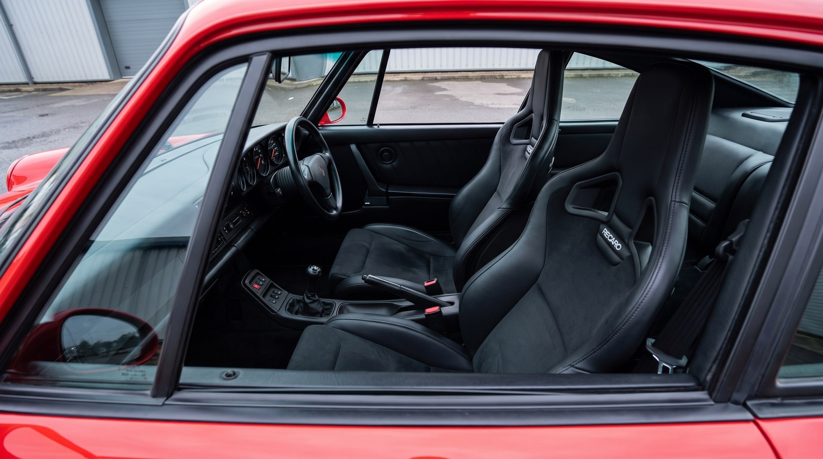 Looking down into the cabin of a red Porsche 964 through the open window of a shut door, showing a pair of Recaro Sportster CS reclining seats facing forward in black leather with suede centre panels, the Recaro wordmark on the shoulder bolster and the classic five dial 911 dashboard ahead of them