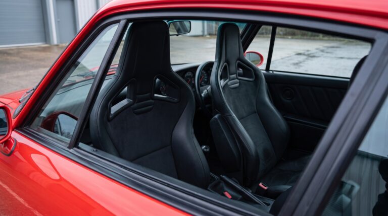 Looking down into the cabin of a red Porsche 964 through the open window of a shut door, showing a pair of Recaro Sportster CS reclining seats in black leather with suede centre panels, their goalpost headrest frames and angled harness slots clearly visible, and the classic five dial 911 dashboard beyond