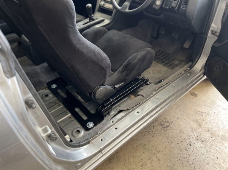 A black suede reclining bucket seat bolted into a right-hand-drive coupe on a flat black steel floor bracket with a double locking sliding runner between the bracket and the seat