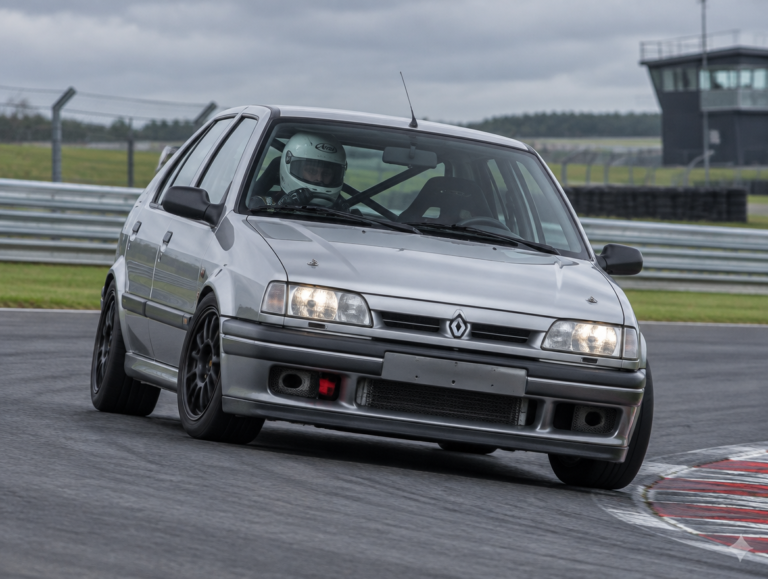 Silver Renault 19 hatchback cornering on a British race circuit with a helmeted driver visible through the windscreen
