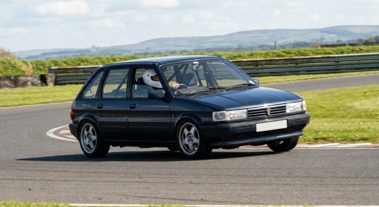 Dark blue Rover Maestro 5-door hatchback cornering on a British race circuit, helmeted driver visible at the wheel