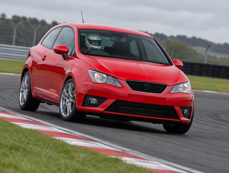 Red Seat hot hatch cornering on a British race circuit with helmeted driver clearly visible through the windscreen
