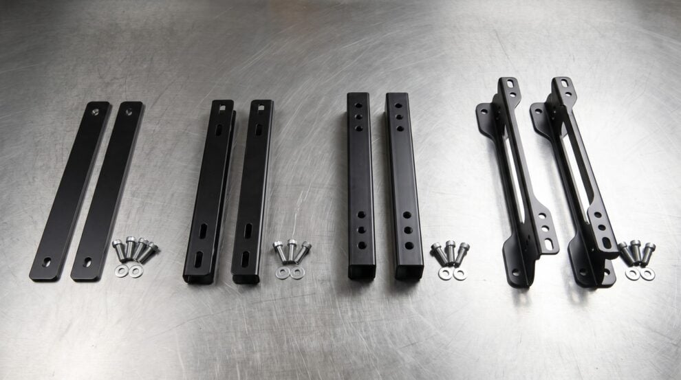 Four kinds of seat spacer laid out on a steel workbench: thin flat strips, one inch channel rails, box section bars with two rows of holes, and tall spacer rails with three holes
