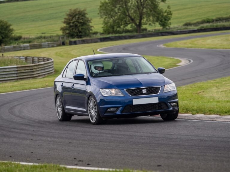 Blue SEAT Toledo Mk3 saloon cornering on a British race circuit with a helmeted driver visible through the windscreen