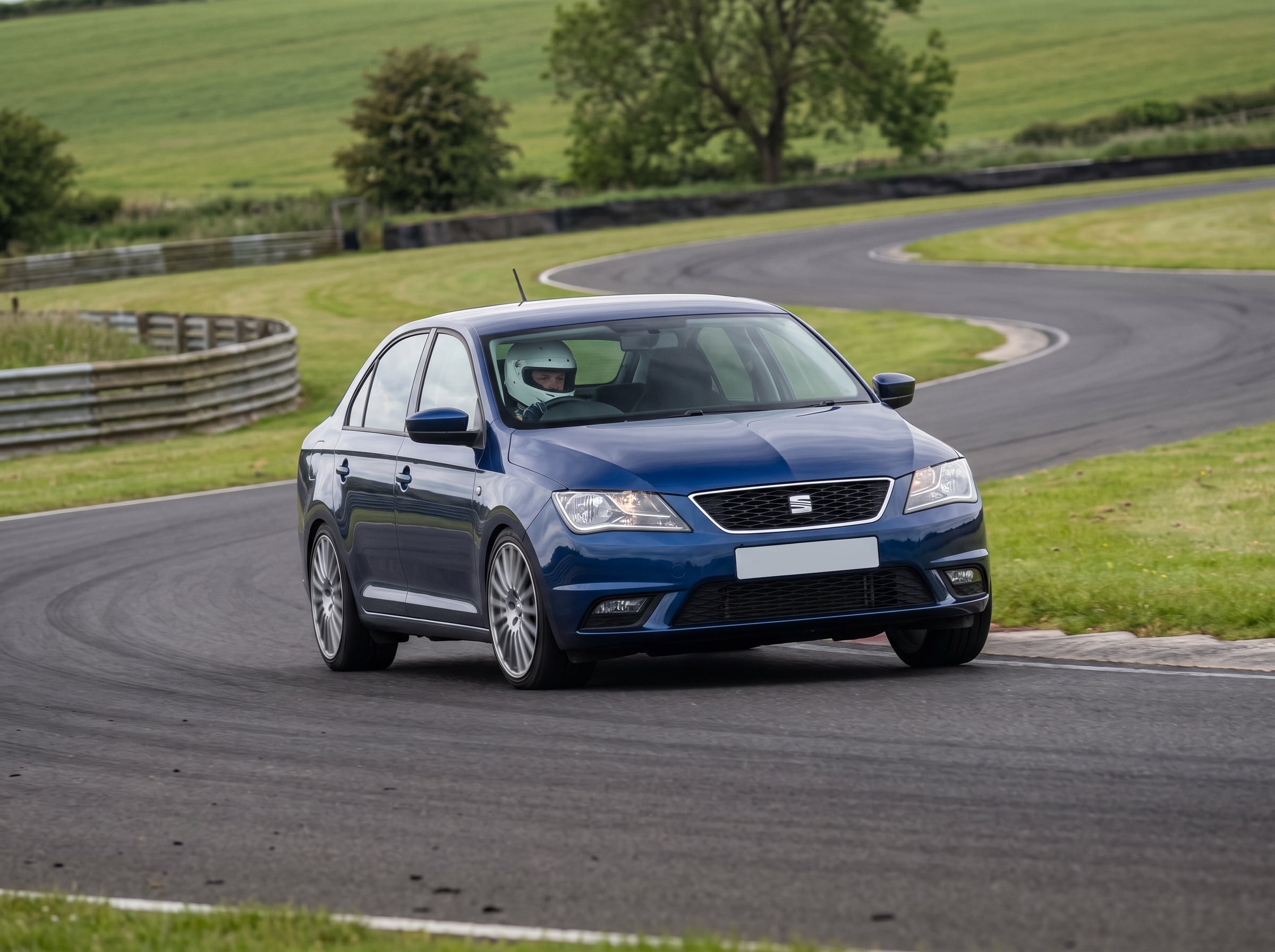 Blue SEAT Toledo Mk3 saloon cornering on a British race circuit with a helmeted driver visible through the windscreen