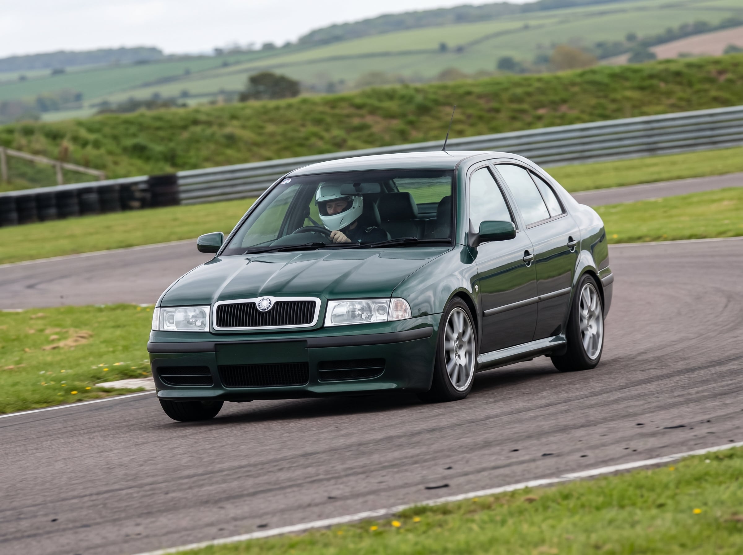 Skoda Octavia Mk1 hatchback cornering on a British race circuit with a helmeted driver visible through the windscreen