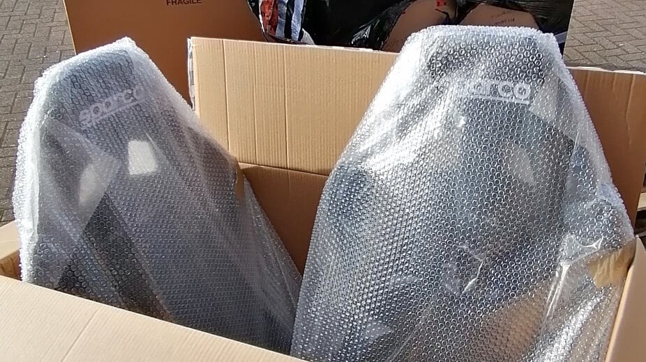 Two bubble-wrapped Sparco bucket seats in an open delivery box in front of a wrapped pallet