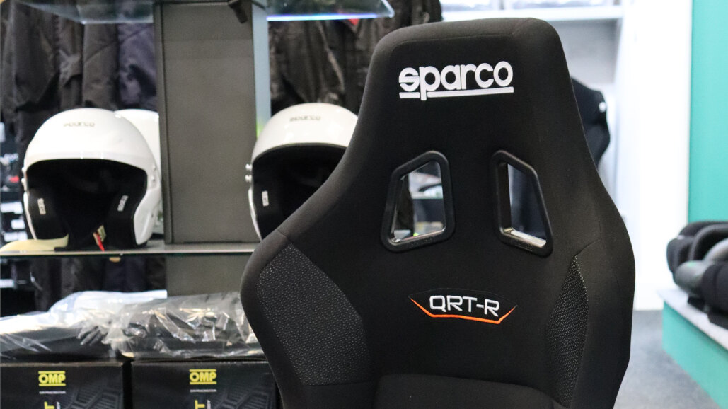 A black Sparco QRT-R bucket seat on the showroom floor with helmets on the shelves behind