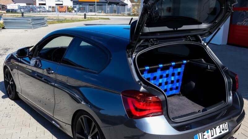 A grey BMW 1 Series with its boot open, showing a Stern Performance strut bar and blue net where the rear seats were