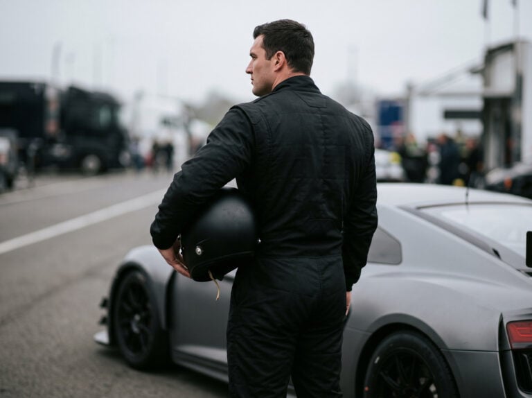 A tall broad shouldered racing driver in a plain black race suit beside a race car