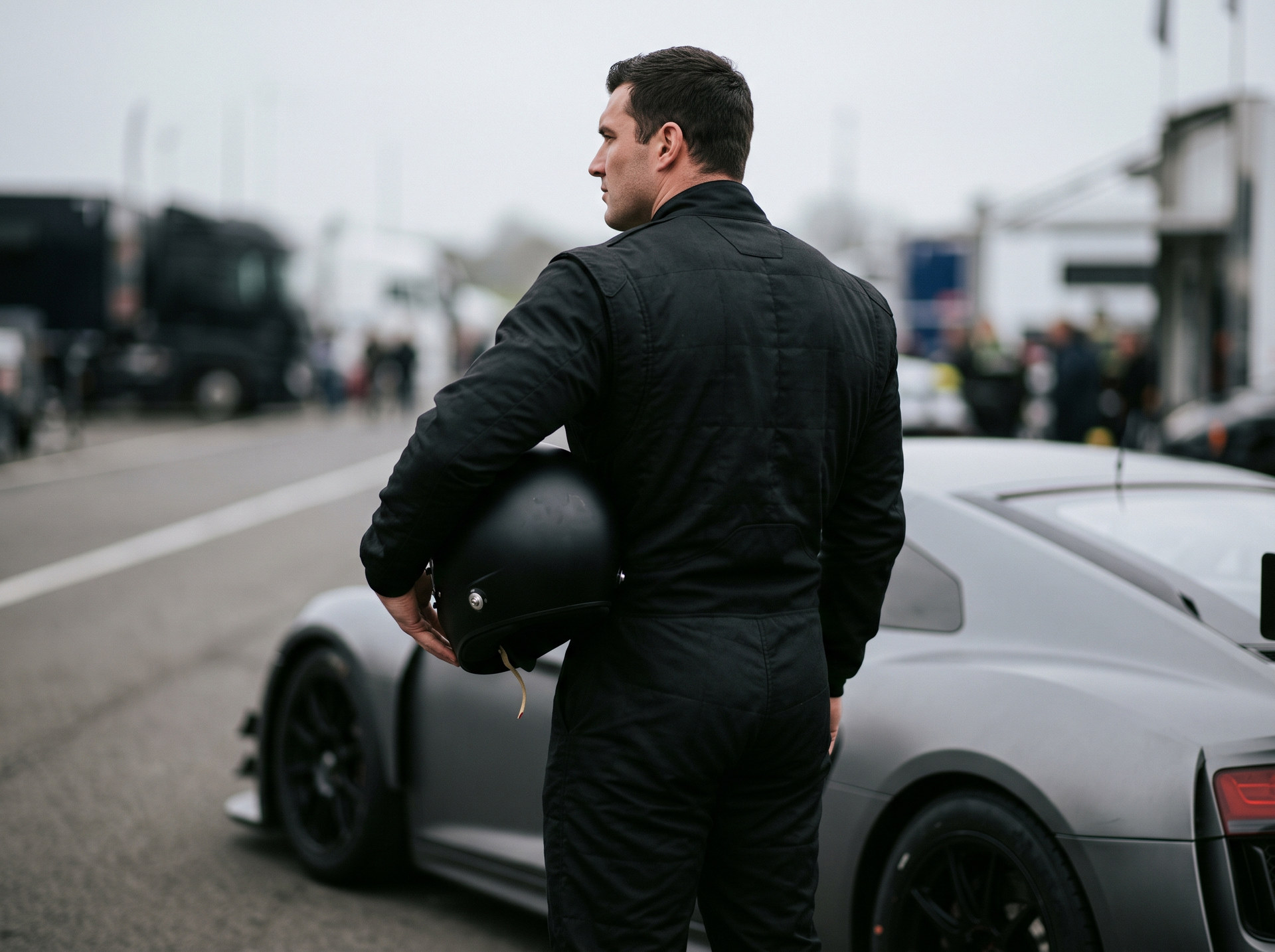 A tall broad shouldered racing driver in a plain black race suit beside a race car