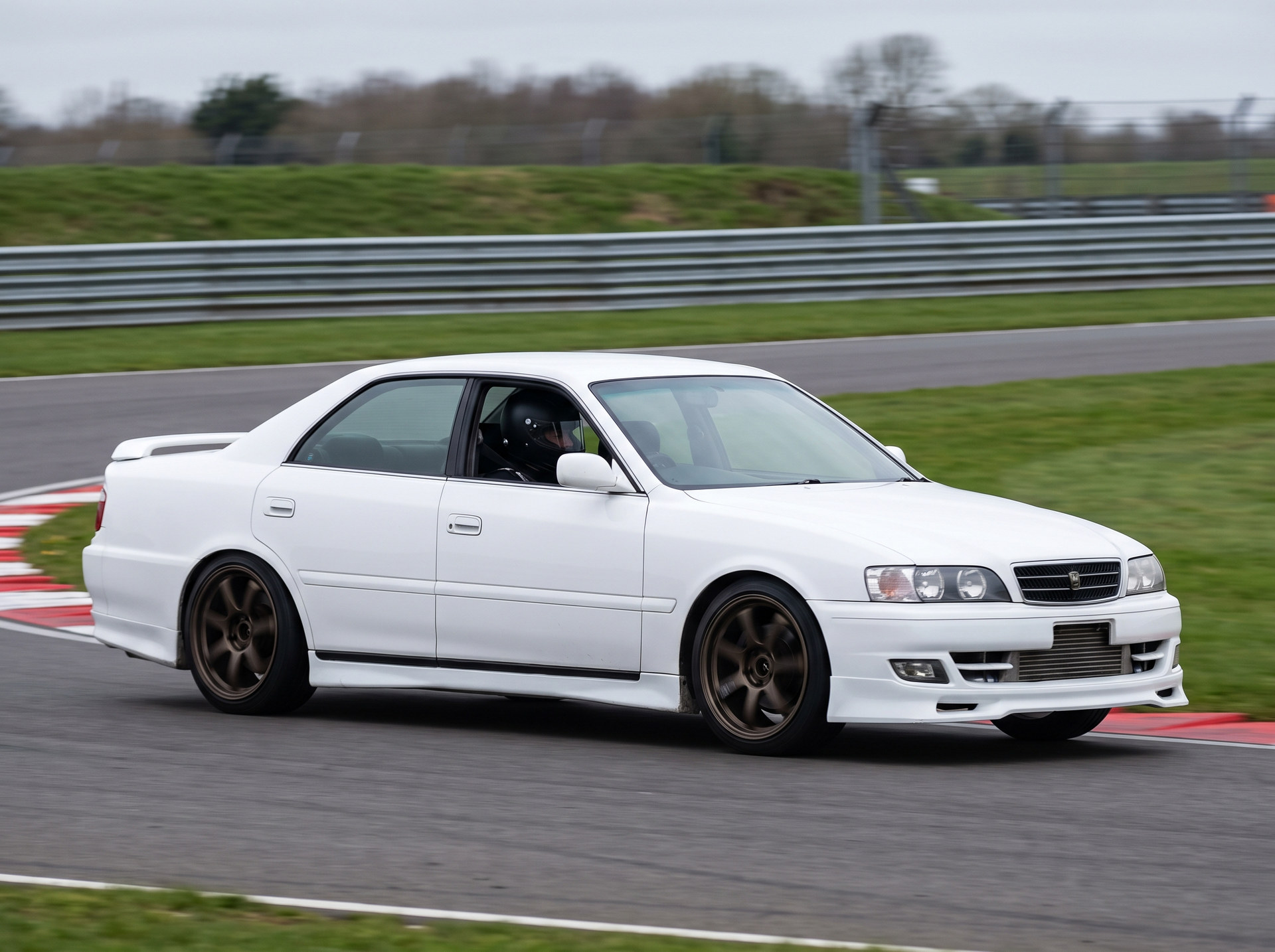 A white Toyota Chaser JZX100 with a helmeted driver cornering on a British race circuit