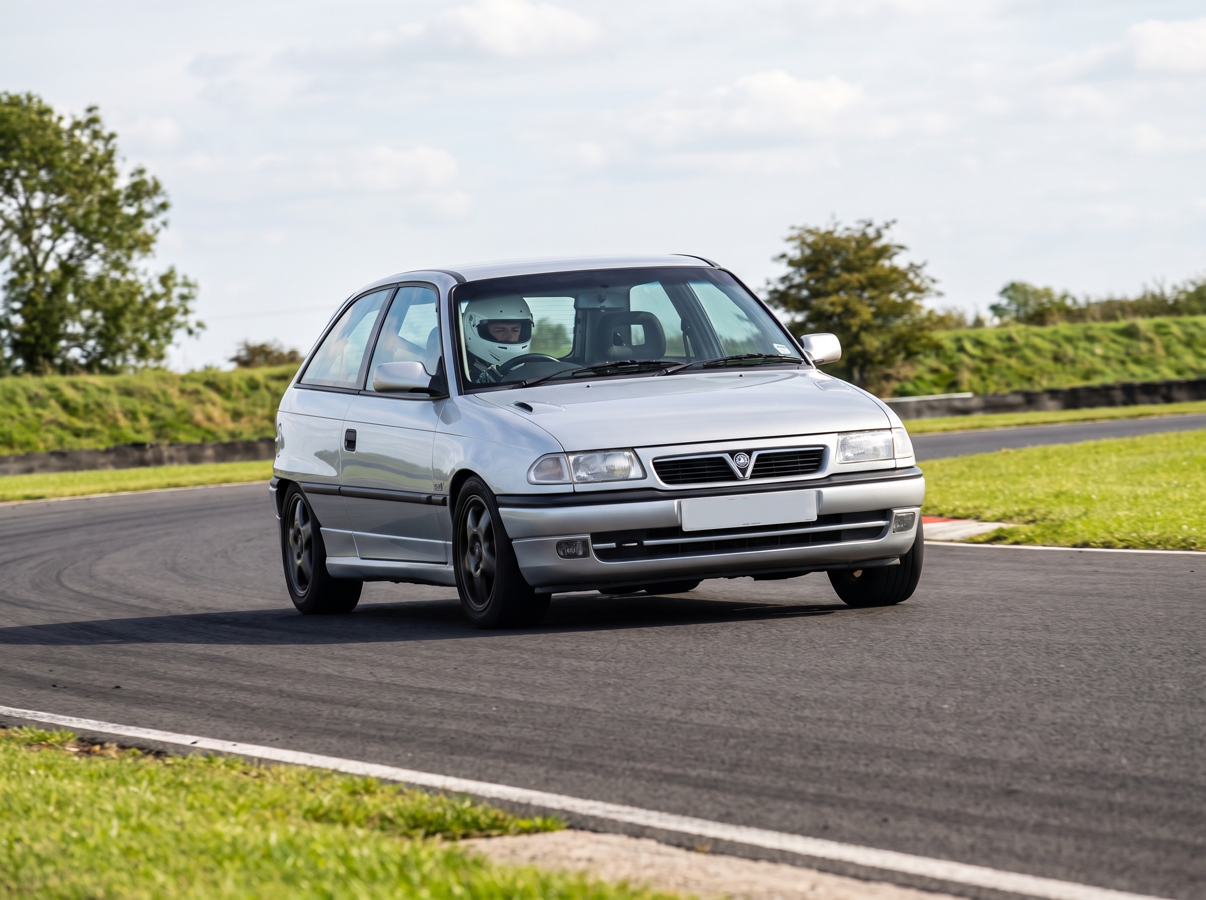Silver Vauxhall Astra Mk3 GSI hot hatch cornering on a British club racing circuit, driver in plain white helmet visible through the windscreen