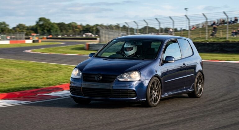 Dark blue Volkswagen Golf Mk5 R32 cornering on a British race circuit, helmeted driver visible through the windscreen