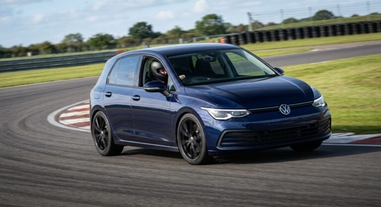 Dark navy blue Volkswagen Golf Mk8 cornering on a British race circuit, helmeted driver visible through the windscreen