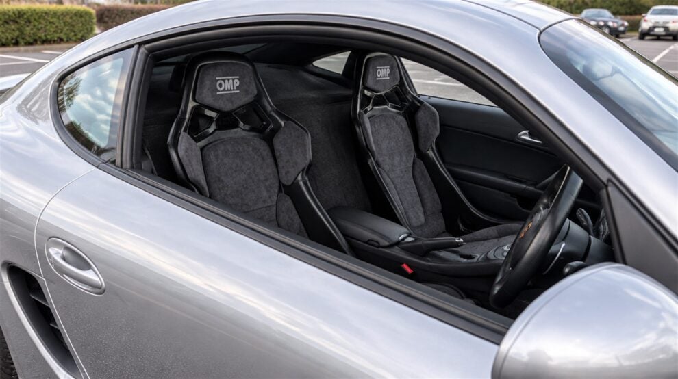 A pair of grey Alcantara OMP RT bucket seats fitted in a silver Porsche Cayman 987, seen through the open driver window with the door closed