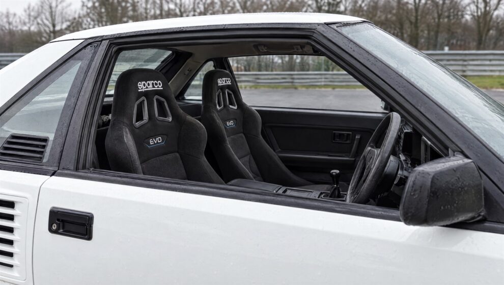 A pair of black Sparco Evo bucket seats with the Sparco logo and EVO badge fitted low in a white Toyota MR2 AW11, seen through the open driver window with the door closed