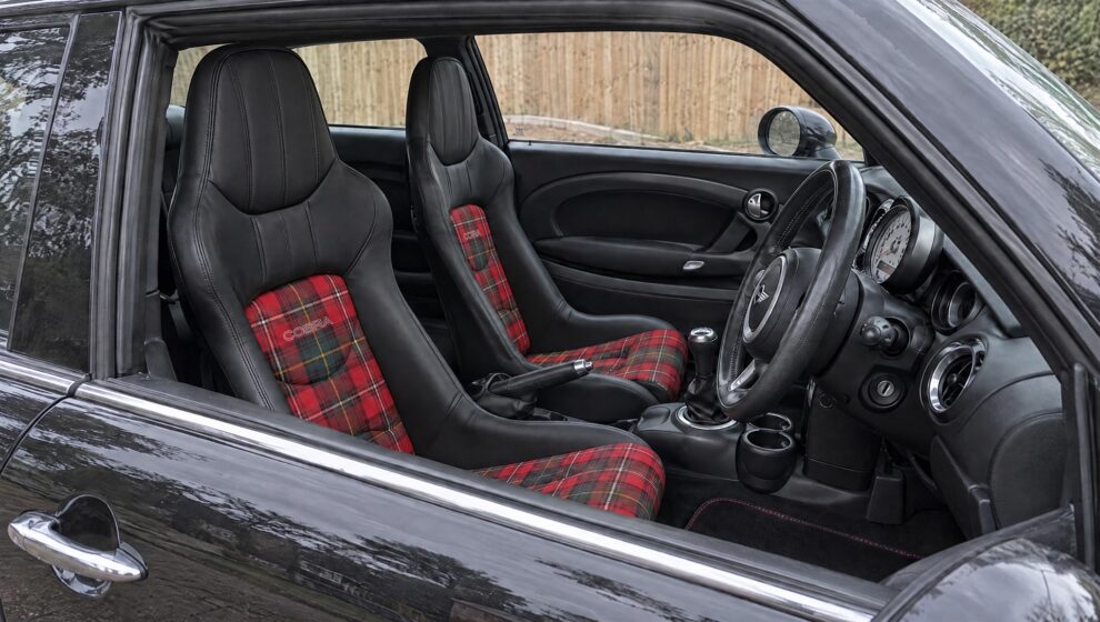 A pair of Cobra Nogaro bucket seats in black leather with a red tartan centre panel and base fitted in the driver and passenger positions of a black Mini R53, seen through the open driver window with the door closed