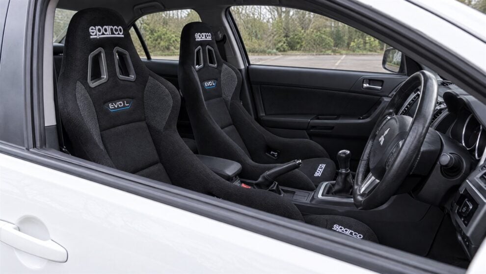 A pair of black Sparco Evo L bucket seats with the Sparco logo and blue EVO L badge fitted in a white Mitsubishi Lancer Evolution X, seen through the open driver window with the door closed