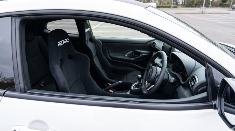 A pair of black velour Recaro Pole Position bucket seats fitted in a white Toyota GR Yaris, seen through the open driver window with the door closed