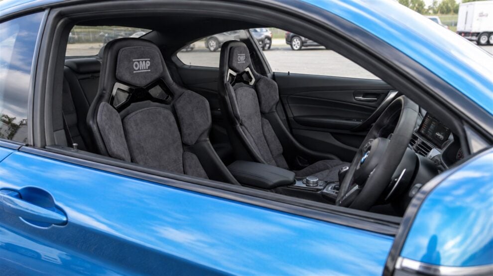 A pair of grey Alcantara OMP RT bucket seats fitted in a blue BMW M2, seen through the open driver window with the door closed