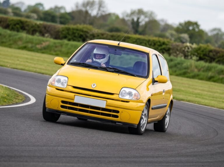 Renault Clio Mk2 Phase 1 cornering at speed on a British race circuit, helmeted driver visible at the wheel