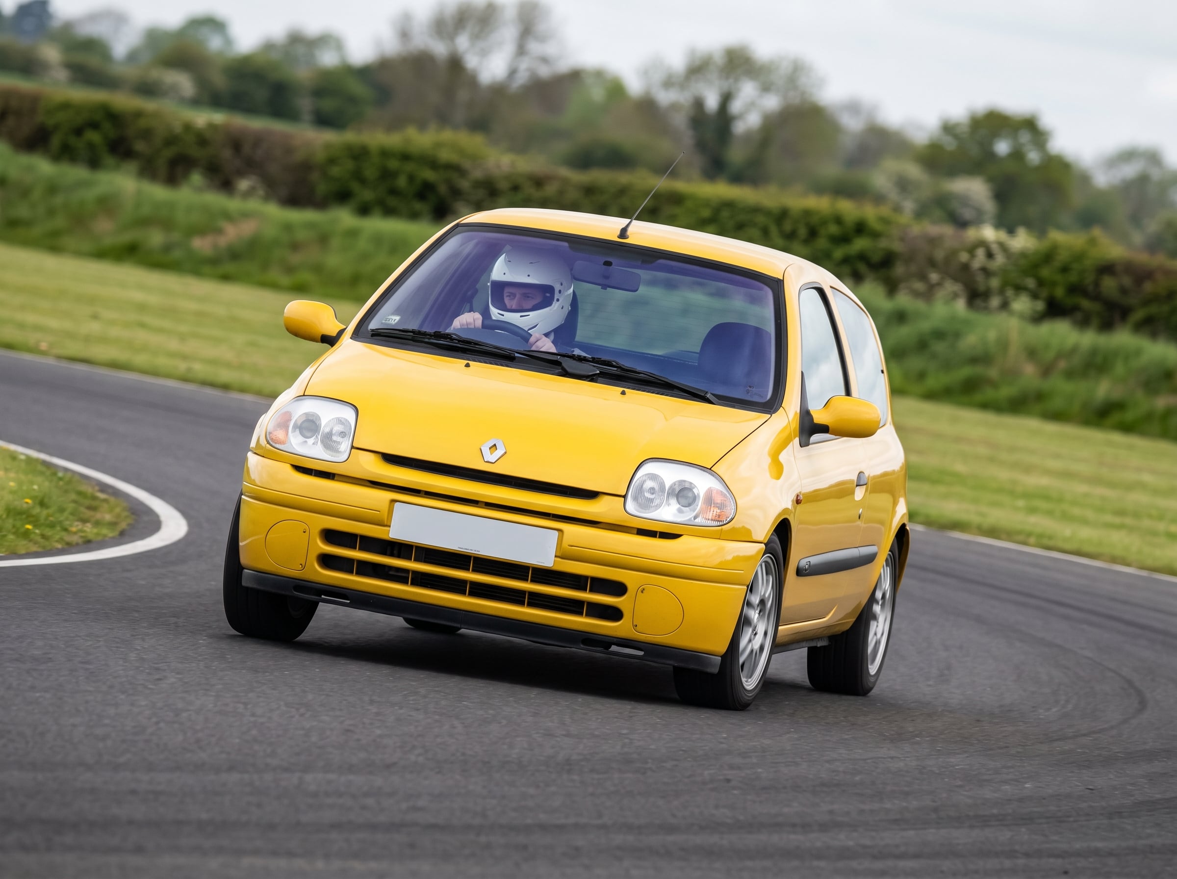 Renault Clio Mk2 Phase 1 cornering on a British race circuit, helmeted driver visible at the wheel